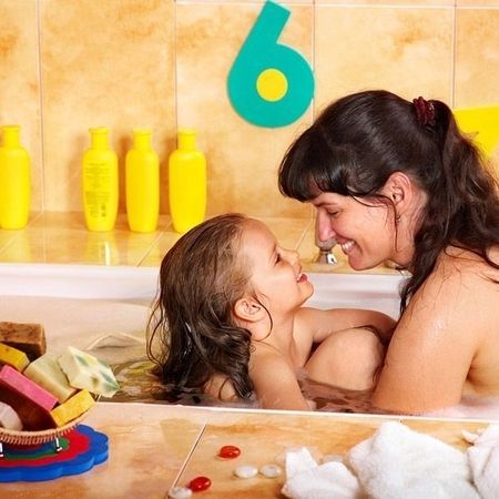 Mutter und Kind sitzen gemeinsam in einer mit Wasser gefüllten Badewanne. Beide schauen sich lächelnd an, die Stimmung ist innig und vertraut. Im Hintergrund stehen mehrere gelbe Shampoo-Flaschen, an der Wand sind bunte Schaumstoffzahlen befestigt. Im Vordergrund liegt Seife in verschiedenen Farben auf einer Seifenablage.