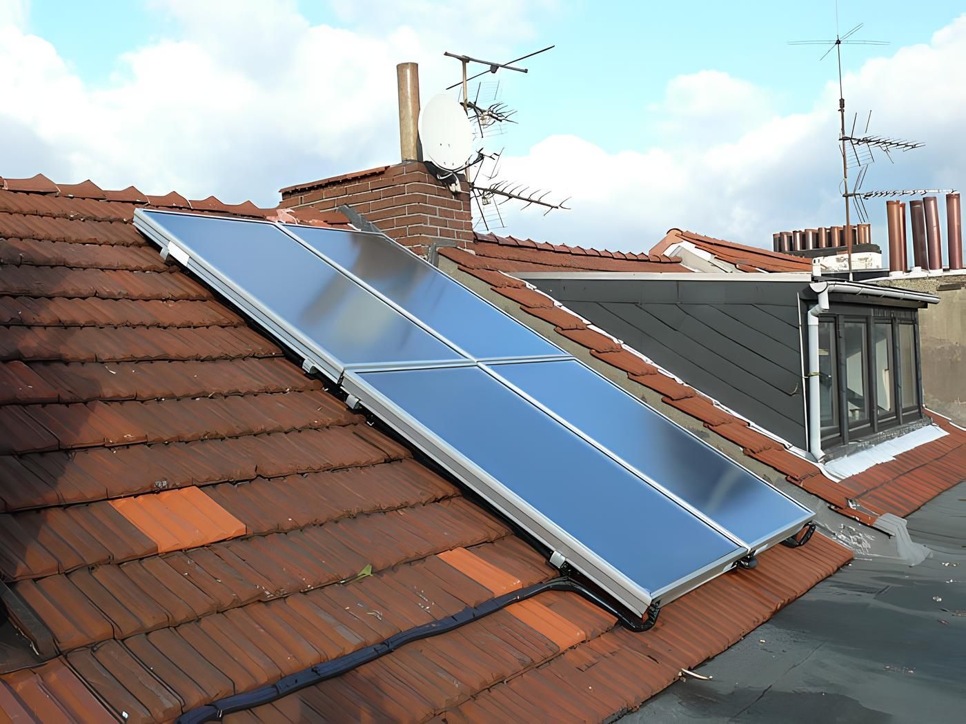 Auf einem geneigten roten Ziegeldach ist ein rechteckiger Solarflachkollektor installiert, daneben befinden sich mehrere Antennen und ein Schornstein.
